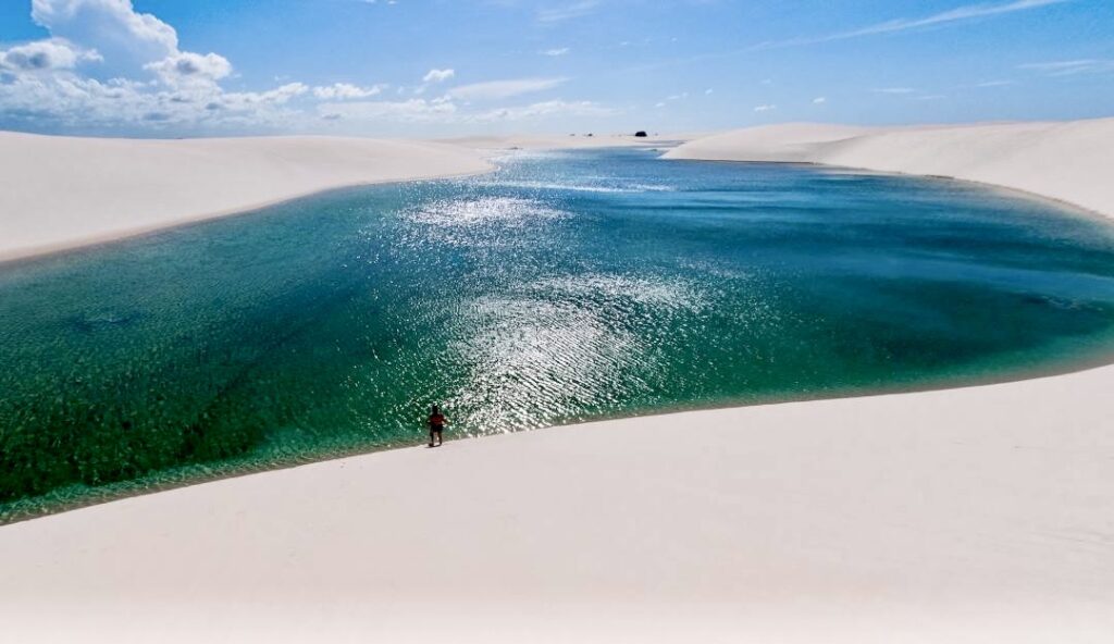 A person standing atop a sand dune in Lençóis Maranhenses, with a stunning green lagoon in the background, capturing the beauty of the national park's unique landscape.