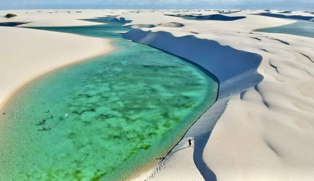 A picturesque view of a towering white sand dune beside a shimmering emerald-green lagoon in Lençóis Maranhenses National Park.