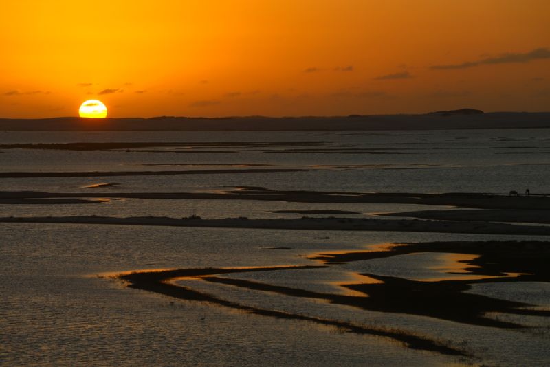 The sun sets over the vast dunes and lagoons of Lençóis Maranhenses, painting the sky in shades of orange, pink, and purple.