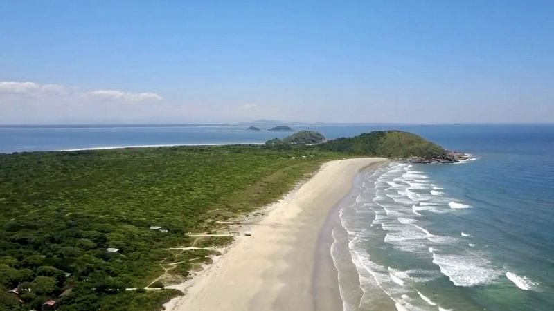 Waves crash onto the golden shores of Praia do Mar de Fora, a wild and breathtaking beach on Ilha do Mel, surrounded by lush green hills.