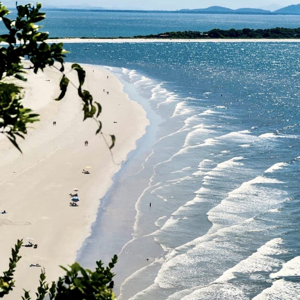 A stunning aerial view of a golden sandy beach on Ilha do Mel, Brazil, framed by lush green hills and the sparkling blue ocean, offering a glimpse of untouched paradise.