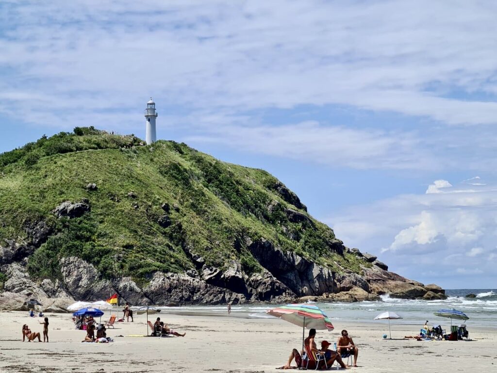 Golden sands and rolling waves of Praia do Farol on Ilha do Mel, with the iconic Conchas Lighthouse standing proudly atop the hill in the background.