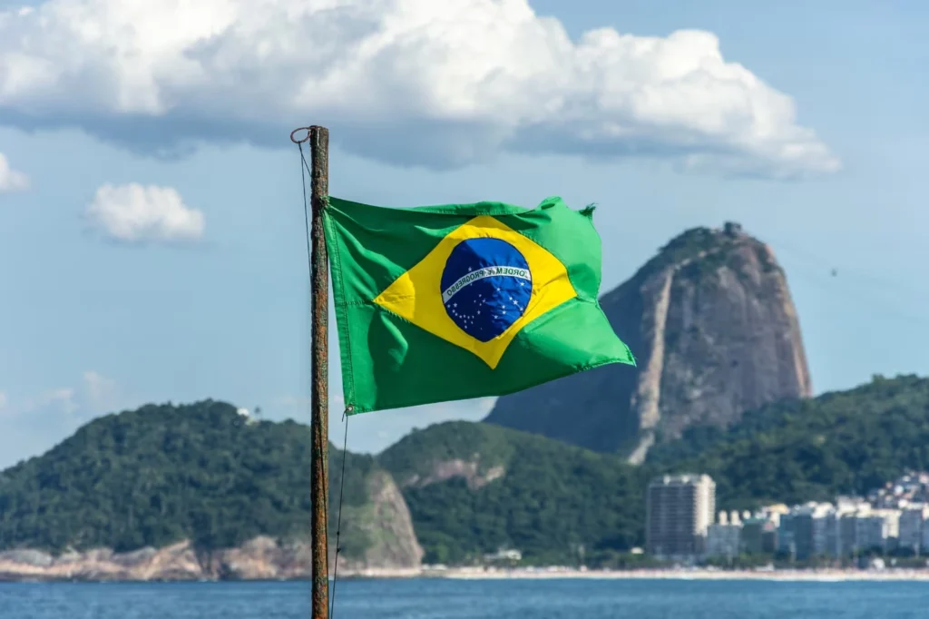 The Brazilian flag waving in the foreground with the majestic Sugarloaf Mountain in the background, symbolizing the nation's beauty and heritage.
