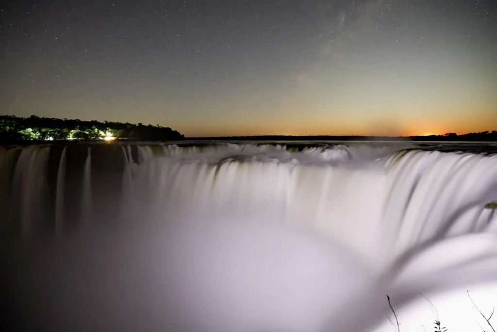 Night view of Iguazu Falls illuminated by moonlight, with mist rising and the surrounding forest in shadow